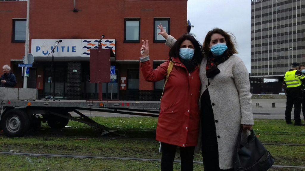 Fatima Faid op de foto met een andere aanwezige op het protest. Ze dragen beide een blauw wegwerpmondkapje en ze maken met hun vingers het vredesteken. Op de achtergrond het politiebureau Marconiplein.