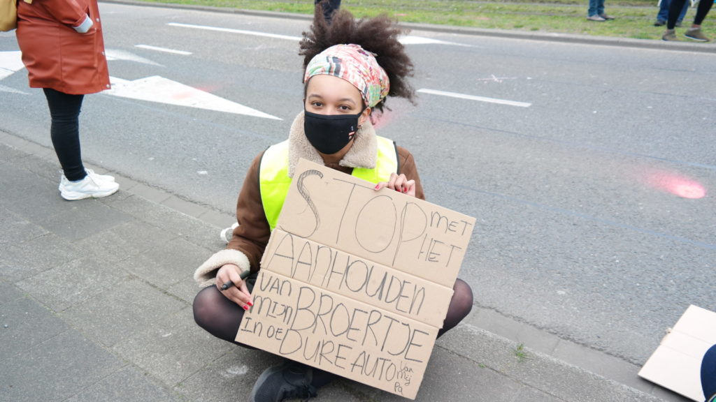 Een demonstrant met een kleurige doek in het haar en een zwart mondkapje zit in kleermakerszit op de stoep met een bord: "Stop met het aanhouden van mijn broertje in de dure auto van mijn pa".