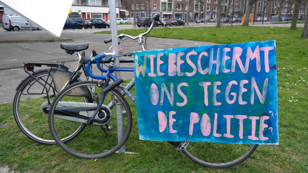 Twee fietsen tegen een paal en een groot blauw bord met de tekst "Wie beschermt ons tegen de politie?".