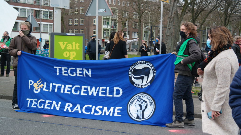 Groot blauw spandoek met AFA Den Haag logo en de tekst "Tegen politiegeweld - tegen racisme". In de letter o van 'politiegeweld' zit het logo van de politie verwerkt.