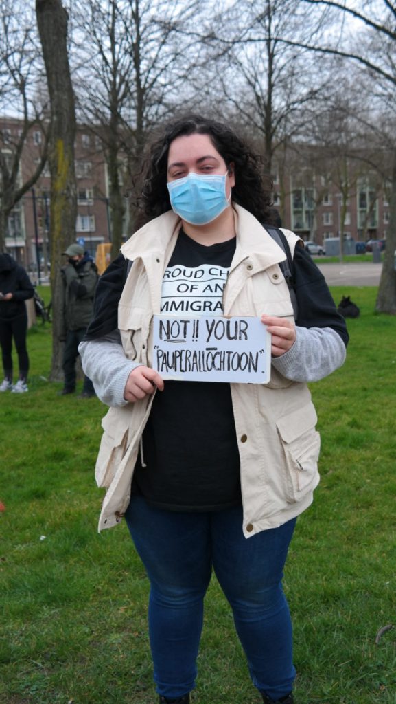 Een persoon met lange zwarte haren en een beige jas draagt een bordje met de tekst "Not your pauperallochtoon".