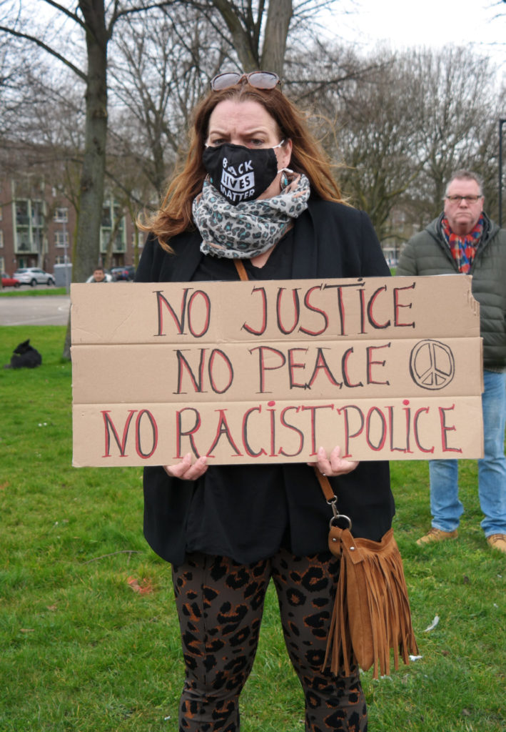 Een ernstig kijkend persoon met lang rossig haar draagt een bord met de tekst "No justice, no peace, no racist police" en een vredesteken.