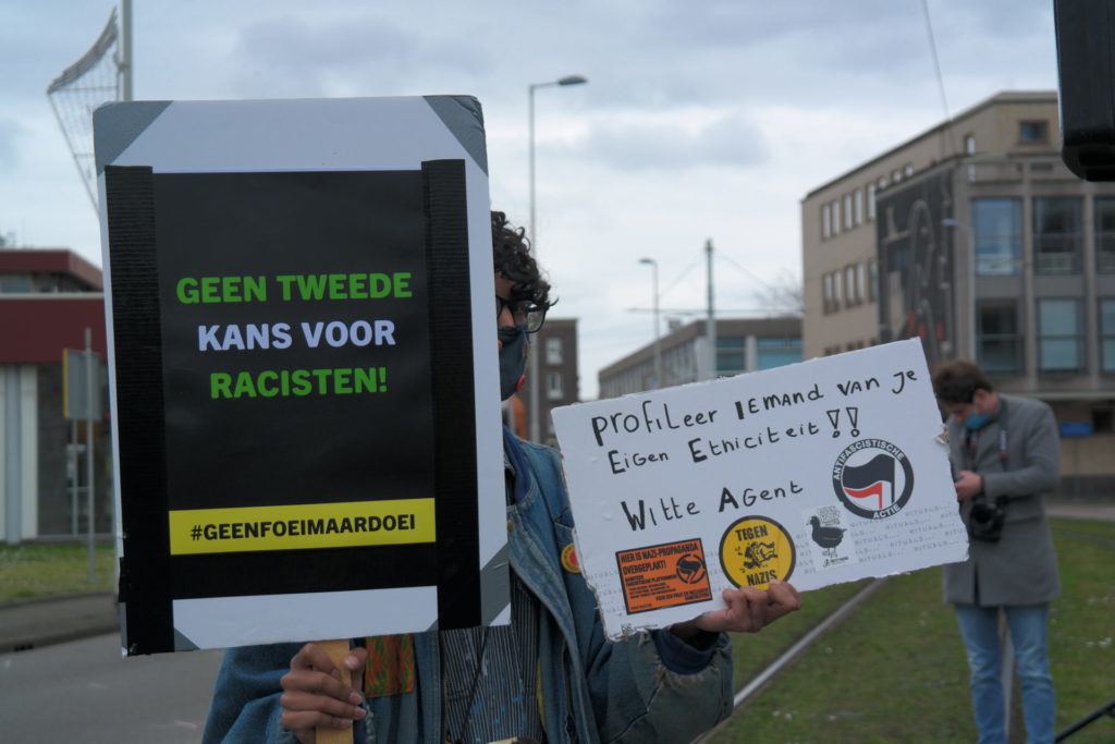 Een demonstrant met kort donker haar draagt twee borden:

"Geen tweede kans voor racisten! #GeenFoeiMaarDoei"

en

"Profileer iemand van je eigen etniciteit, witte agent!!"