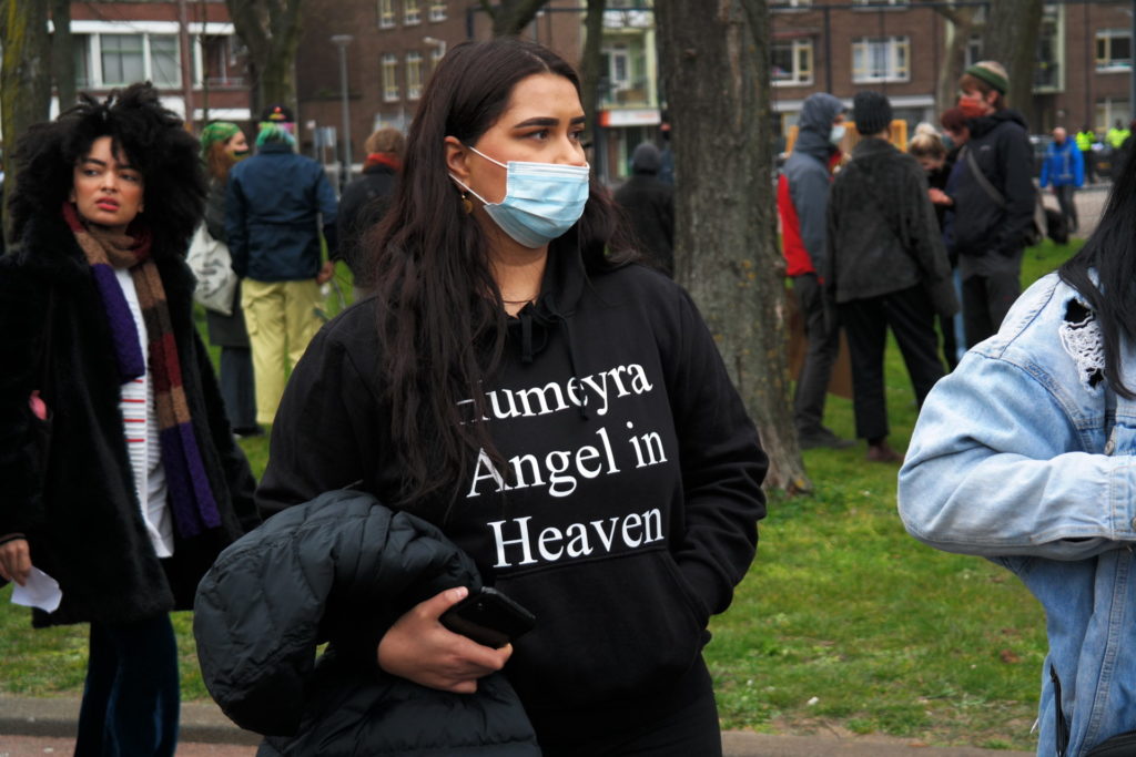 Een persoon met lange zwarte haren en een blauw wegwerpmondkapje op kijkt naar rechts. Ze draagt een zwarte trui met de tekst "Humeyra Angel in Heaven".