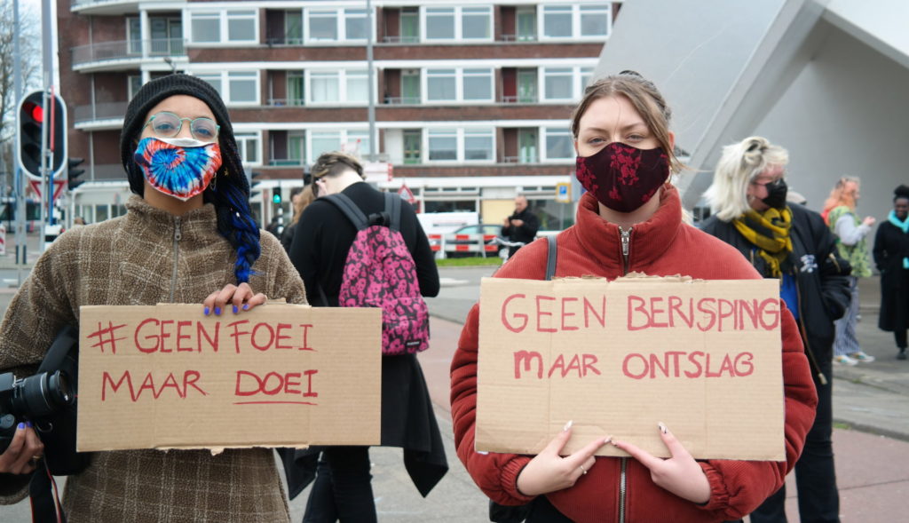 Twee personen met fleurige mondkapjes en borden met de teksten:

"#GeenFoeiMaarDoei" 

en

"Geen berisping maar ontslag".