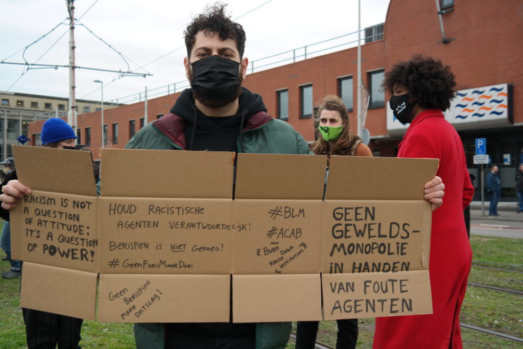 Een persoon met kort haar en een zwart mondkapje over hun baard houd een uitgeklapte doos als protestbord vast. Er staat veel tekst op: "Racism is not a question of attitude: it's a question of power!" / "Houd racistische agenten verantwoordelijk! Berispen is NIET genoeg! #GeenFoeiMaarDoei" / "Geen berisping maar ontslag!" / "#BLM #ACAB Burn down racist institutions" / Geen geweldsmonopolie in handen van foute agenten".