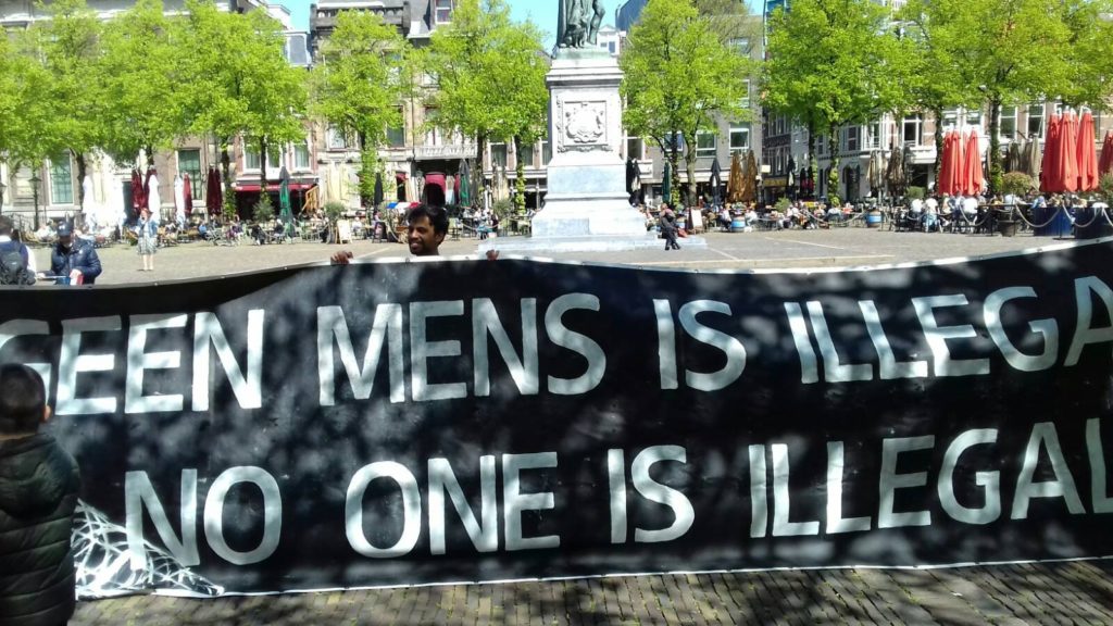 Enorm spandoek op een zonnig plein, de tekst luidt: "Geen mens is illegaal / No one is illegal".