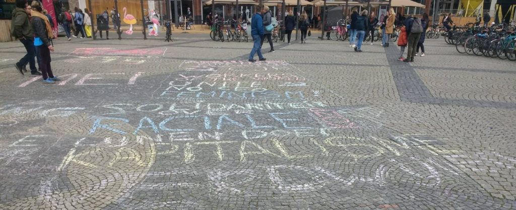 Allemaal verschillende krijtteksten op een plein bestraat met kinderkopjes. De teksten zijn niet heel goed leesbaar door de hoek van de foto.