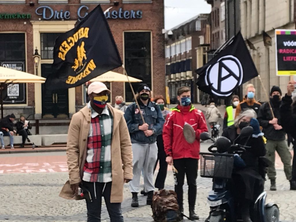 Groepje demonstranten in Groningen. Grote, zwarte anarchisme-vlag, 'Refugees welcome'-vlag, persoon in een scootmobiel.