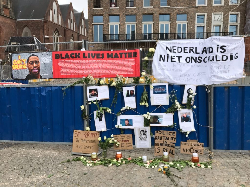 Een bouwhek met daarop foto's van Nederlandse zwarte personen / personen van kleur die zijn omgekomen door politiegeweld, waaronder Mitch Henriques en Tomy Holten. Er hangt een spandoek met de tekst "Nederland is niet onschuldig" en de namen van slachtoffers van politiegeweld, waaronder Rishi Chandrikasing en Ihsan Gürz.