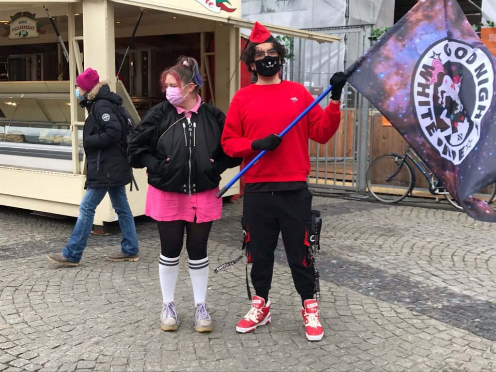 Een jonge persoon in een rood-zwarte outfit met een zwarte kattenmondkap en een roodzwart hoedje op draagt een eenhoorn-vlag met de tekst "Good night white pride". Naast hem staat iemand in een korte roze rok en een zwart kort jack.