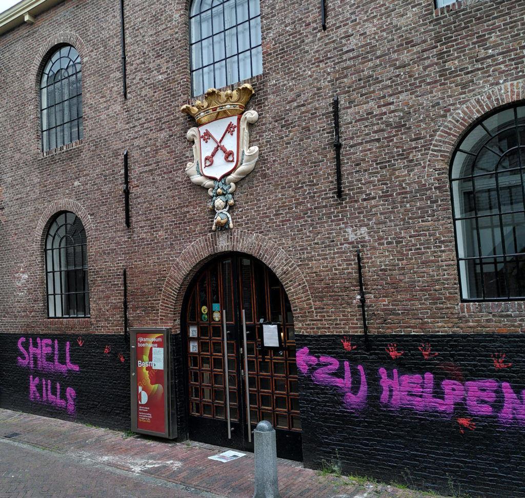 De muur van een oud gebouw met een toegangspoort met daarboven een stadswapen. Naast de poort staat in knalpaars "Shell kills - zij helpen" met ernaast een pijl richting de poort.