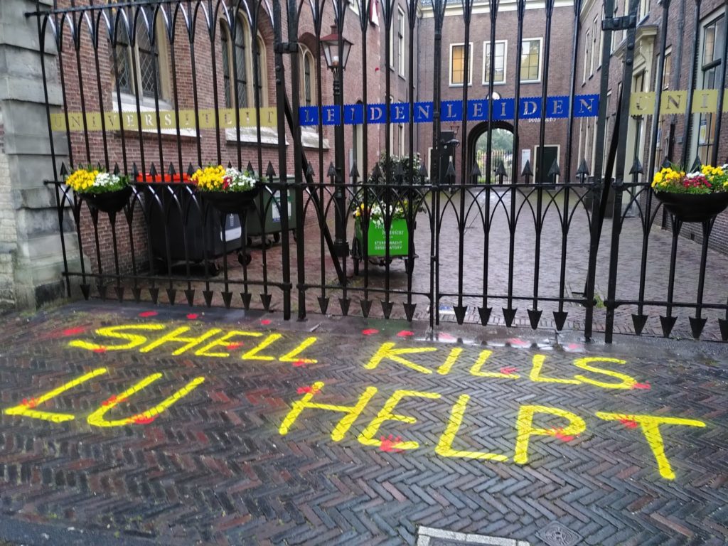 Felgele tekst op de bestrating voor de universiteit: 'Shell kills, LU helpt'. De tekst is voorzien van felrode handafdrukken.