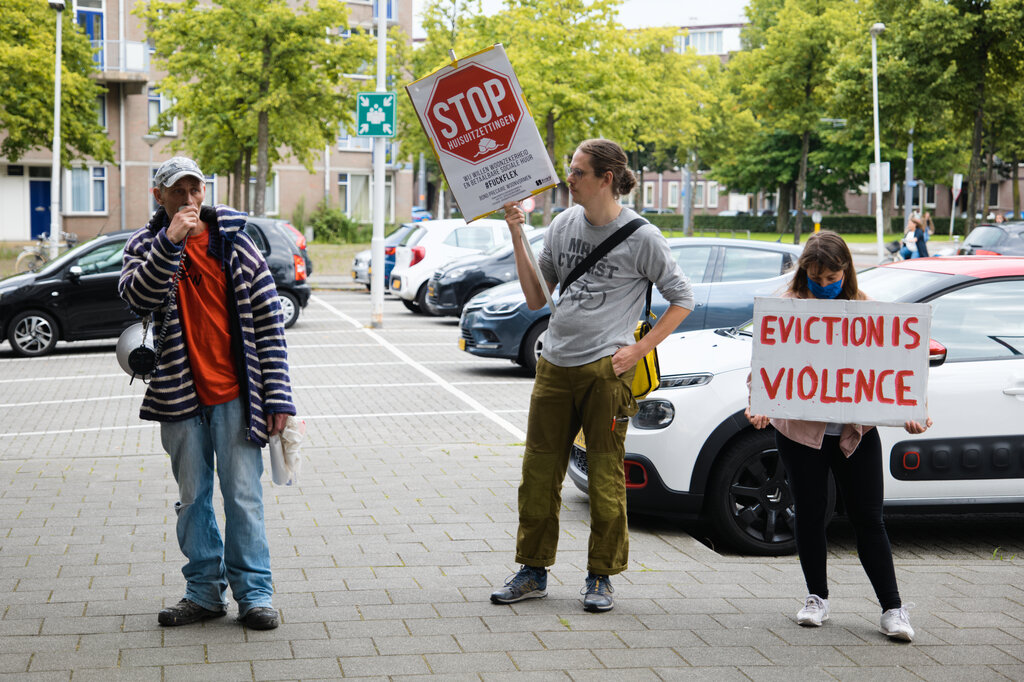 Iemand spreekt de demonstranten toe via een megafoon. Twee anderen staan ernaast met borden met "Stop huisuitzettingen" en "Eviction is violence".