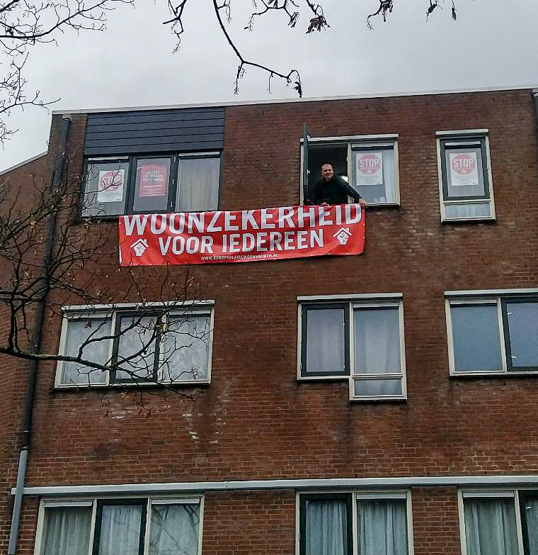 Een man is te zien vanuit het open raam van zijn woning. Hij lacht de kamera toe. Buiten zijn raam hang een rood spandoek met de tekst 'woonzekerheid voor iedereen'. Achter de ramen van zijn woning hangen posters met de tekst 'stop huisuitzettingen'.