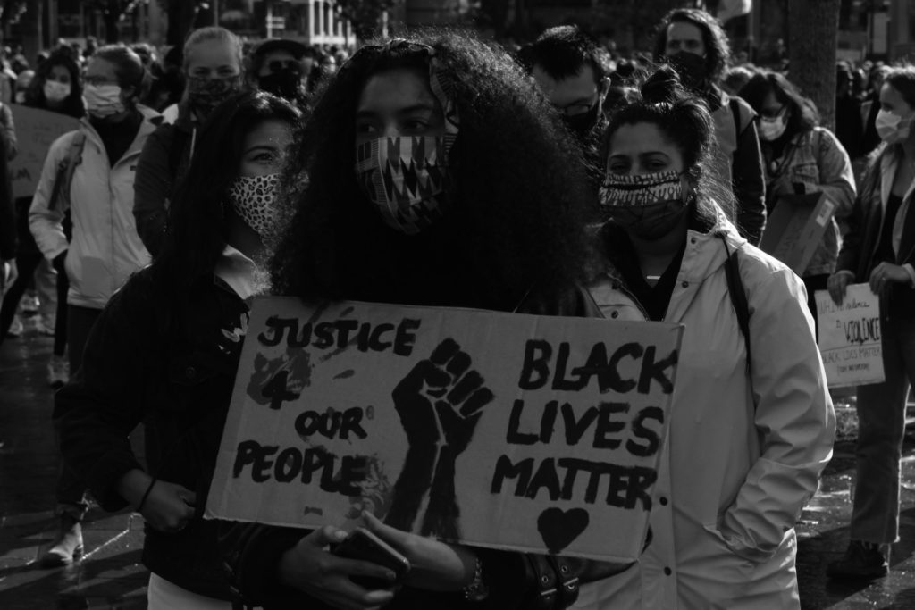 Een persoon met een mondkapje draagt een bord met de tekst "Justice 4 our people - black lives matter".