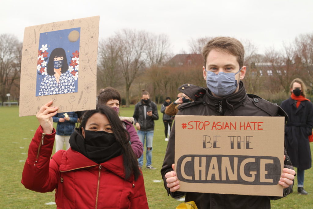 Een Aziatisch persoon met lange haren houdt een bord omhoog met daarop een kleurrijke afbeelding van een Aziatisch persoon met een mondkapje met de tekst "Hate the virus". Naast haar staat een wit persoon met korte haren en een bord met de tekst "Stop Asian hate - be the change".