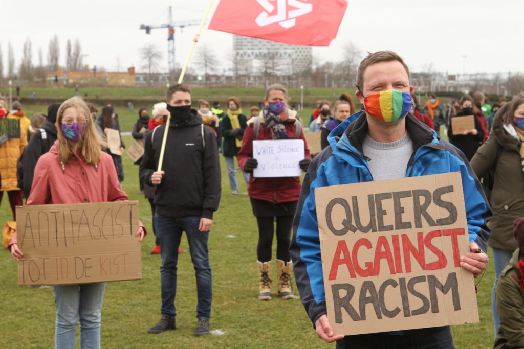 Een persoon met een regenboogmondkapje en een bord met de tekst 'Queers against racism'. Iets verder naar achteren een persoon met een roze jas en een bord met de tekst 'Antifascist tot in de kist'.