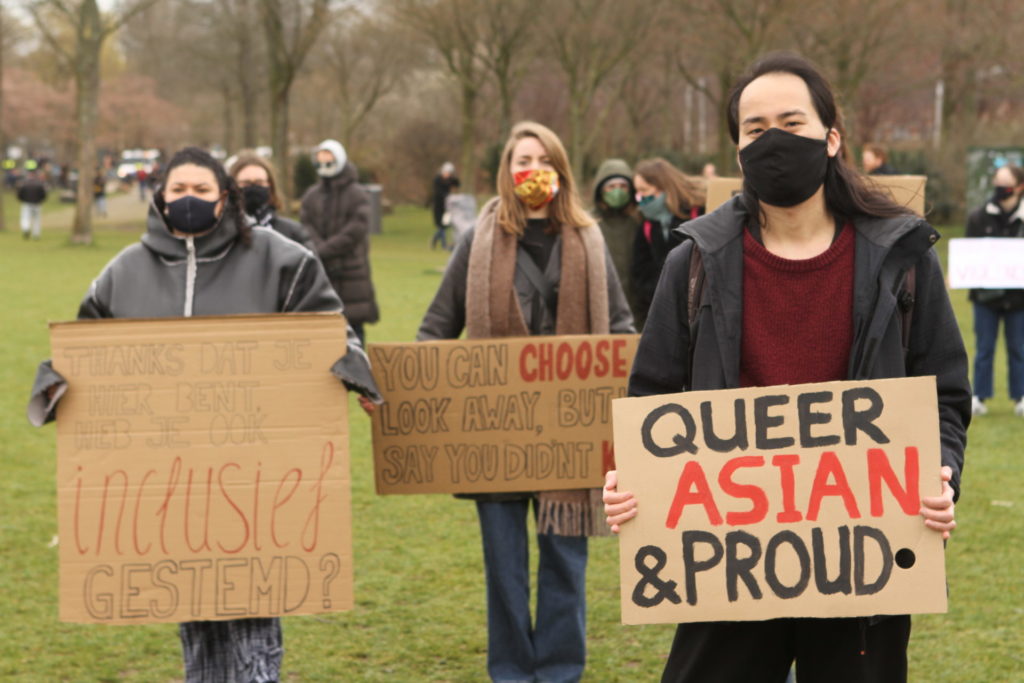 Drie personen poseren met hun borden voor de foto. Twee borden zijn goed leesbaar met de volgende teksten:

"Thanks dat je hier bent, heb je ook inclusief gestemd?"

"Queer, Asian & proud".