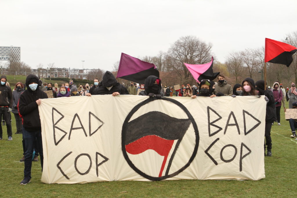 Vier in het zwart gekleden personen met een groot spandoek met antifa-logo met de tekst "Bad cop bad cop".