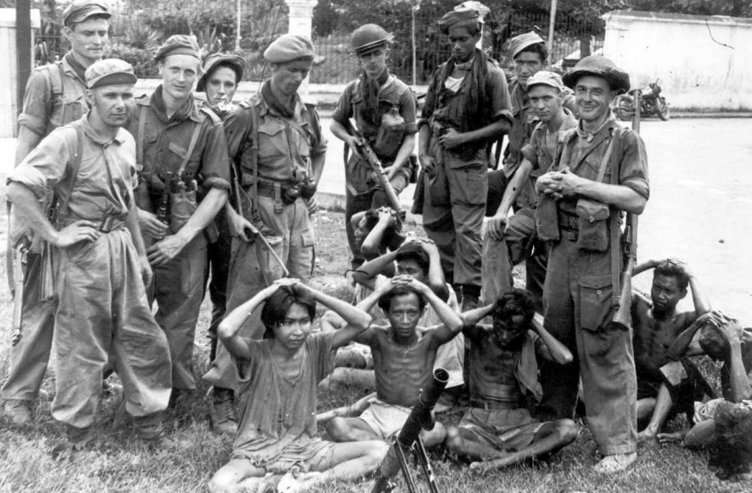 Een zwartwitfoto waarop Nederlandse soldaten een aantal Indonesische gevangenen onder schot houten. De gevangenen zitten allemaal met hun handen op het hoofd. De soldaten staan er opvallend relaxed bij, eentje heeft zelfs een brede grijns op zijn gezicht.