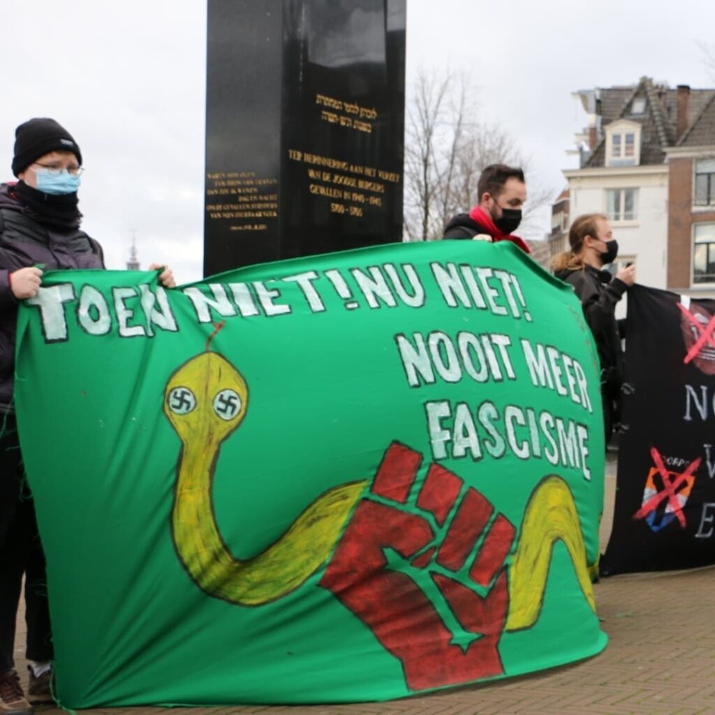 Mensen op een demonstratie dragen een spandoek. Op het spandoek is een rode vuist te zien, en die vuist grijpt een gele slang met hakenkruizen als ogen. Ook is op het spandoek de tekst 'toen niet, nu niet, nooit meer fascisme' te lezen.