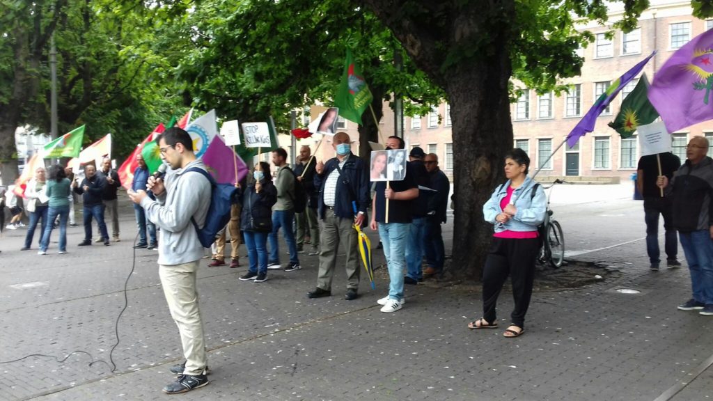 De demonstranten staan met allerlei verschillende vlaggen en protestborden onder een grote boom.