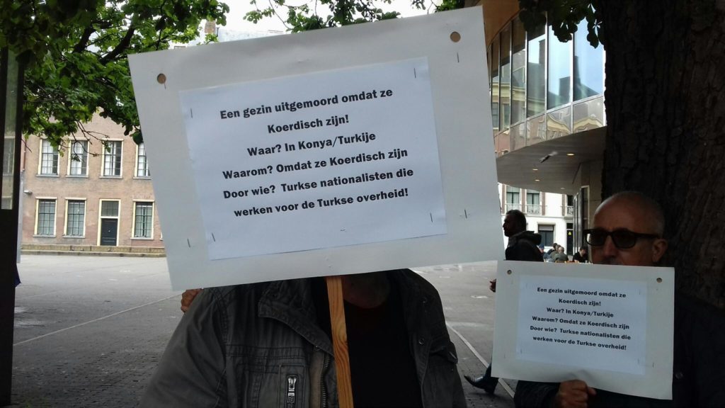Een demonstrant gaat schuil achter een bord met de tekst "Een gezin uitgemoord omdat ze Koerdisch zijn! Waar? In Konya/Turkije. Waarom? Omdat ze Koerdisch zijn. Door wie? Turkse nationalisten die werken voor de Turkse overheid!"