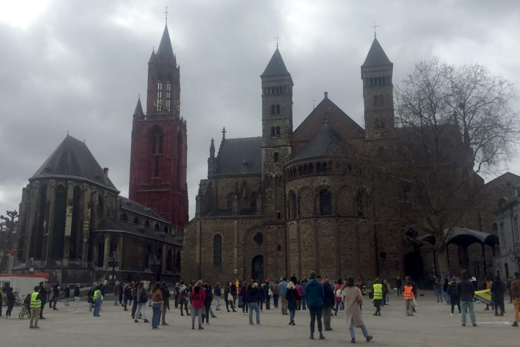 Het Vrijthof in Maastricht, guur weer. Je ziet de kerk en de basiliek met daarvoor tientallen demonstranten.