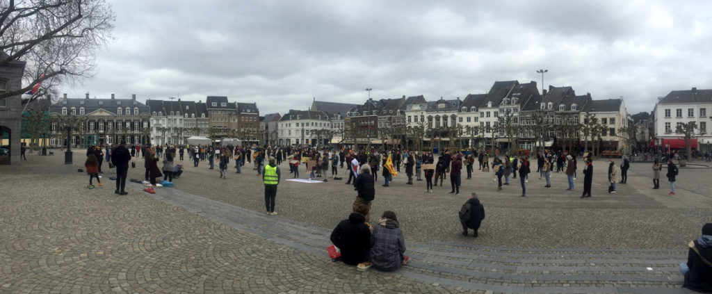 Overzichtsfoto van de demonstratie op het Vrijthof. Op de voorgrond trappetjes waar enkele mensen op zitten. Daarachter allemaal staande demonstranten.