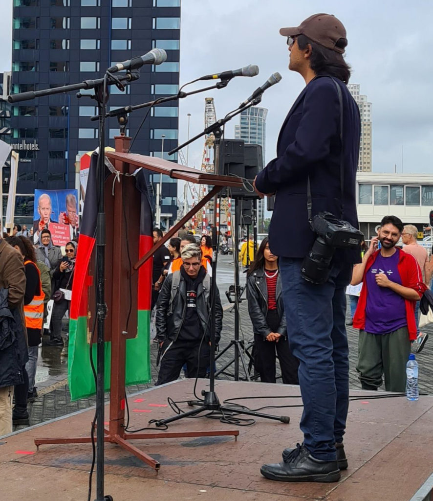 Massoud spreekt de demonstranten toe vanachter een lessenaar waar een Afghaanse vlag aan hangt. Massoud draagt een bruin petje, een blauw jasje en een spijkerbroek.