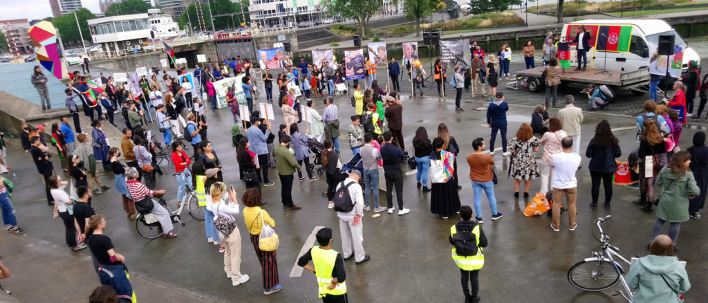 Overzicht van de demo van bovenaf gezien. Het podium is een aanhangwagen. Erachter een bus met Afghaanse vlaggen en protestborden.