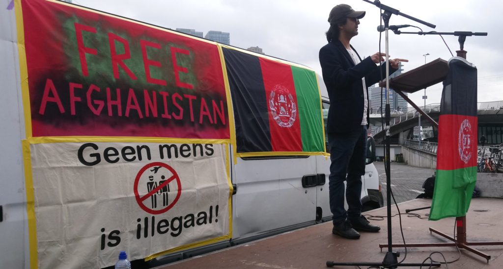 Massoud op het podium met achter zich de bus met spandoeken, teksten 'Free Afghanistan' en 'Geen mens is illegaal!'.