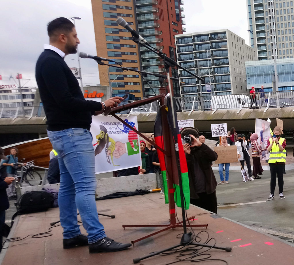 Milad op het podium. Hij draagt een donkerblauwe trui met daaronder een overhemd en jeans. Hij heeft zijn haar achterover gekamd en hij draagt een kort baardje.