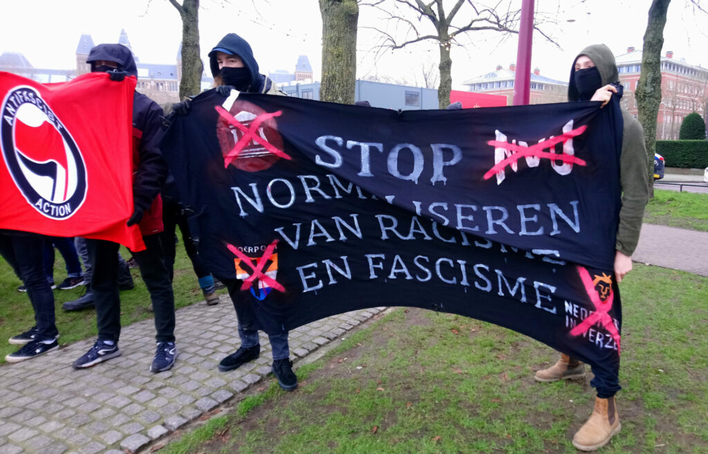 Enkele anti-fascistische demonstranten. Twee dragen een spandoek met de tekst 'stop normaliseren van racisme en fascisme'.