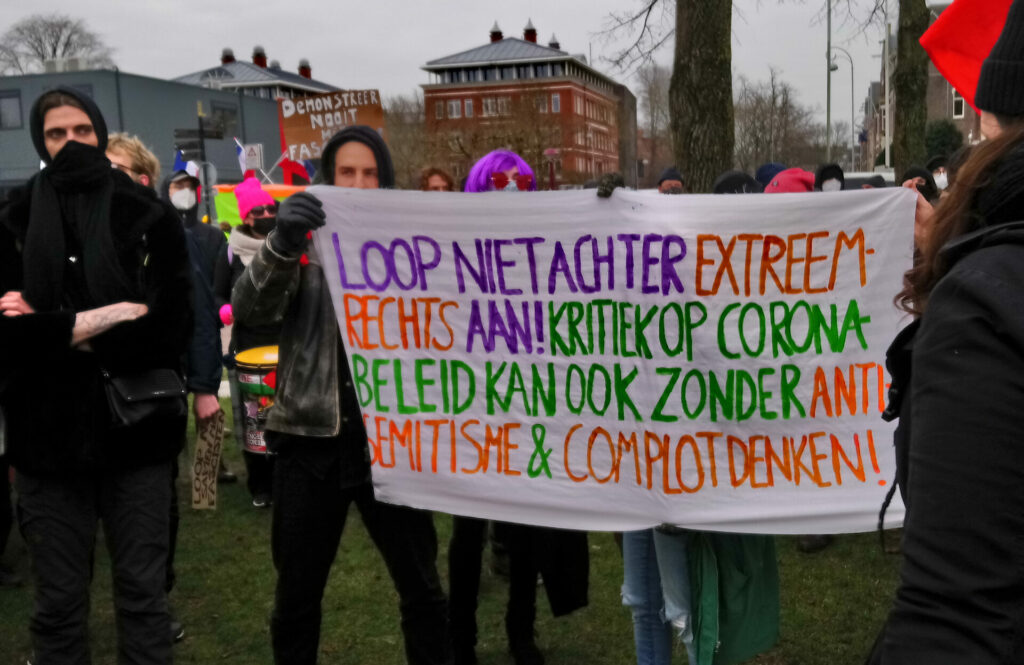 Een groep antifascistische demonstranten. Twee houden een spandoek op met de tekst 'loop niet achter extreem-rechts aan! kritiek op corona beleid kan ook zonder anti-semitisme en complotdenken'.