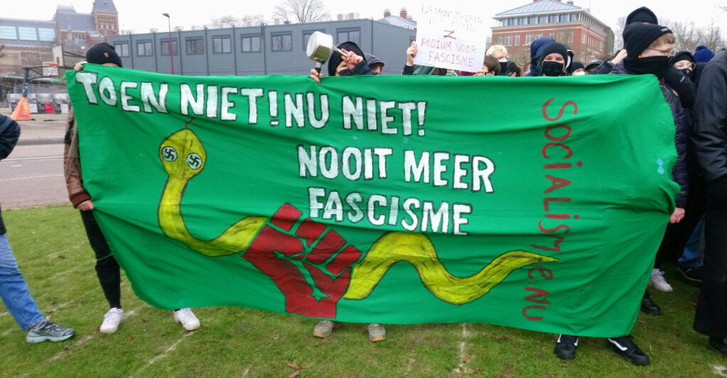 Een groep antifascistische demonstranten. Enkelen houden een spandoek op met een rode vuist die een gele slang grijp; de slang heeft hakenkruizen als ogen. Er staat ook de tekst 'Toen niet! Nu niet! Nooit meer fascisme!'

