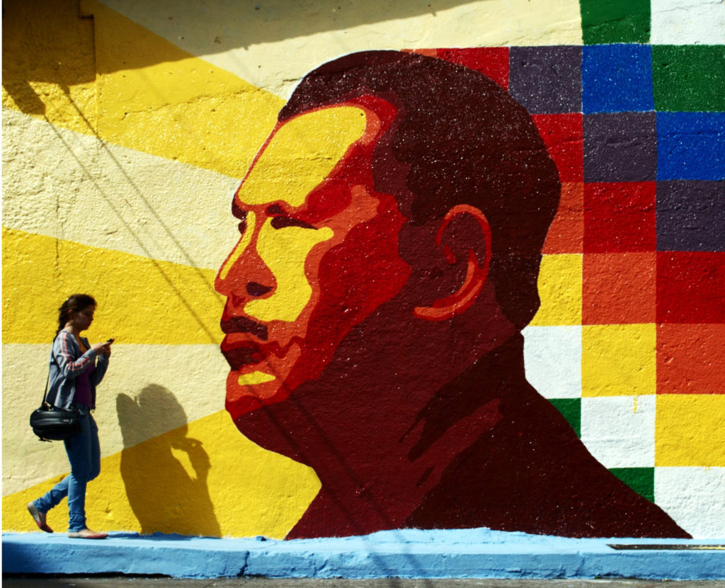 Kleurrijke wandschildering van Hugo Chavez. Een jong iemand loopt er voorbij, kijkend op een smartphone.