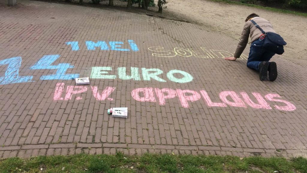Iemand zit geknield op de grond en maakt een krijttekst "1 mei solida-". Eronder staat "14 euro ipv applaus".