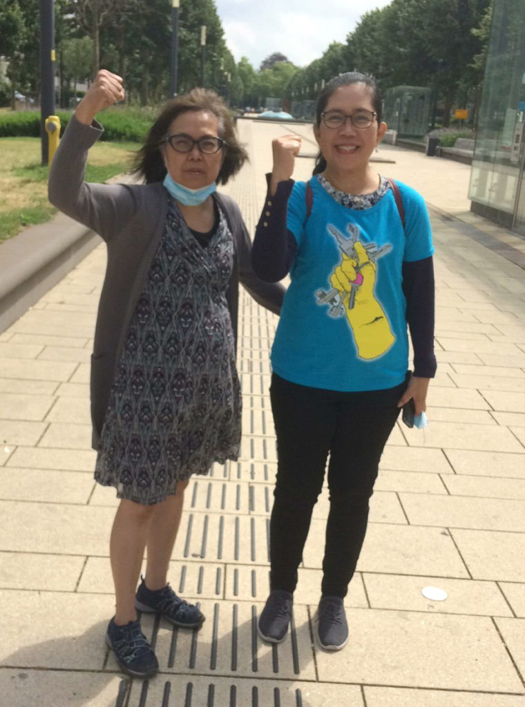 Twee deelnemers aan de demonstratie, beide met de vuisten in de lucht. Eentje draagt een T-shirt met daarop een hand gehuld in een schoonmaakhandschoen die een bos sleutels vasthoudt.