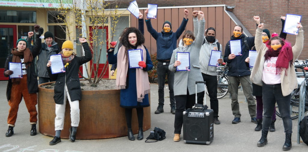 Een groep demonstranten rondom een grote bloembak. De meeste van hen dragen mondkapjes van Voor 14. Ze hebben allemaal een A4 met een tekst in hun handen en ze heffen hun gesloten vuist. Op de grond staat een geluidsinstallatie.