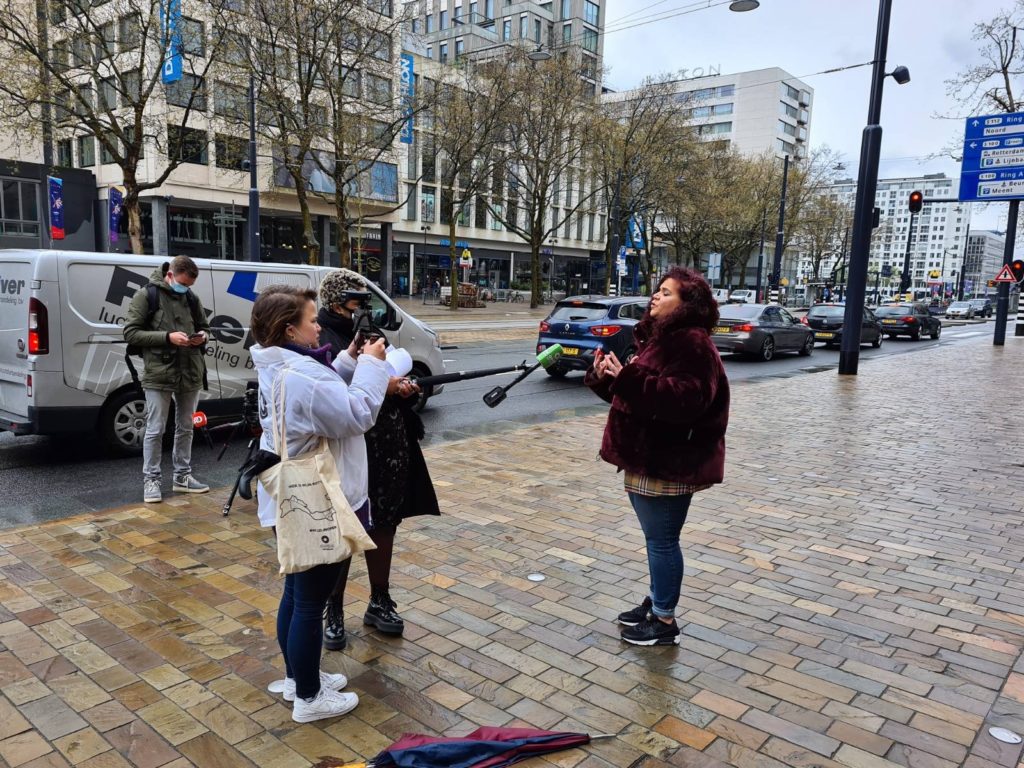 Sandra Salome geeft buiten voor het stadhuis een interview aan de pers. Het is regenachtig weer. Sandra draagt een auberginekleurige fluffy jas van nepbont.