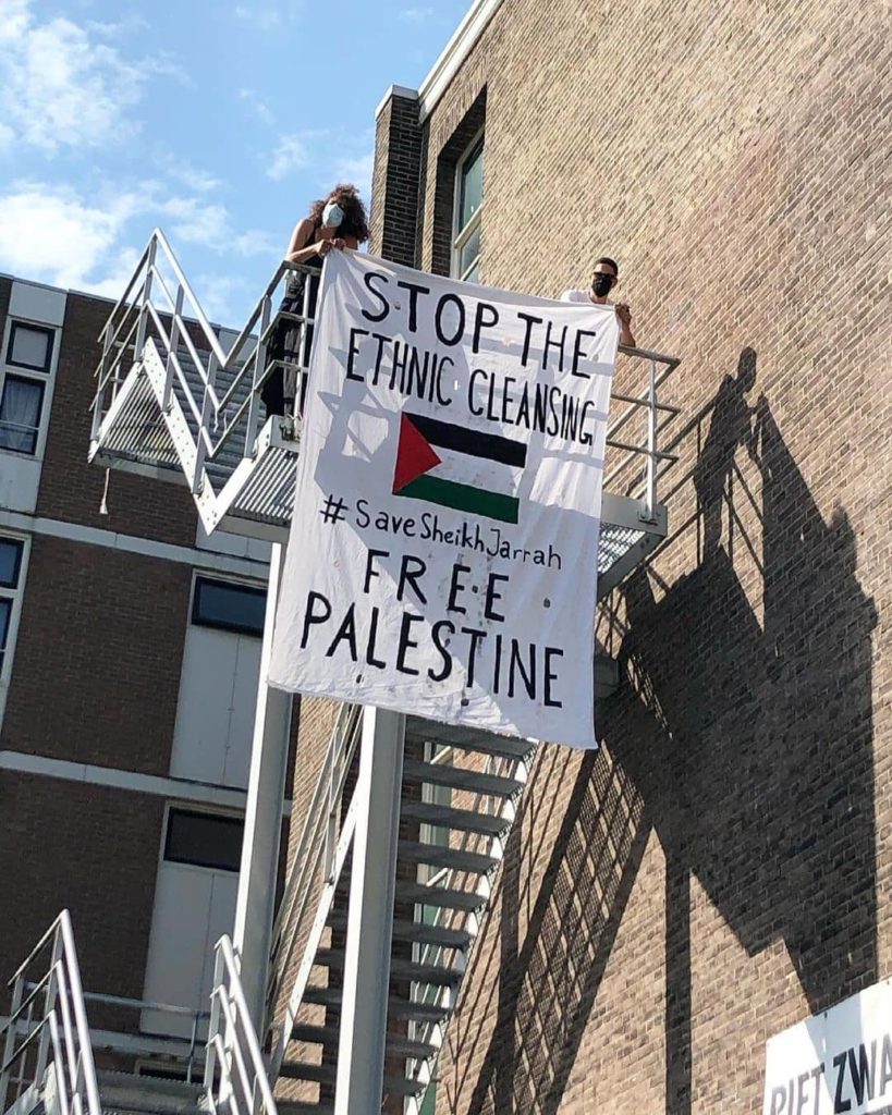 Twee studenten leunen op de reling van de brandtrap en houden het spandoek vast.