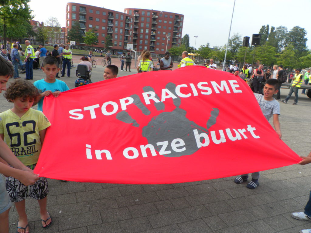 Kinderen met een groot, rood spandoek met daarop een hele grote grijze handafdruk en de tekst "STOP RACISME in onze buurt".