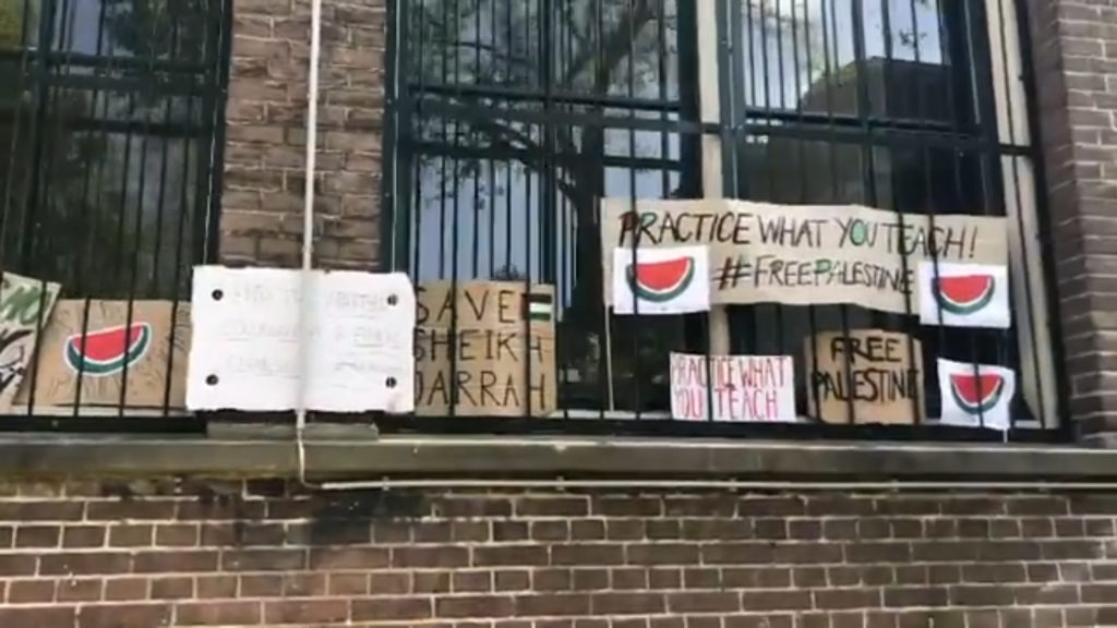 Verschillende protestborden achter een hekwerk bij de ramen van de school. Teksten:
- "Practice what you teach! #FreePalestine"
- "Save Sheikh Jarrah"
- "Free Palestine"

Tevens meerdere tekeningen van meloenen.