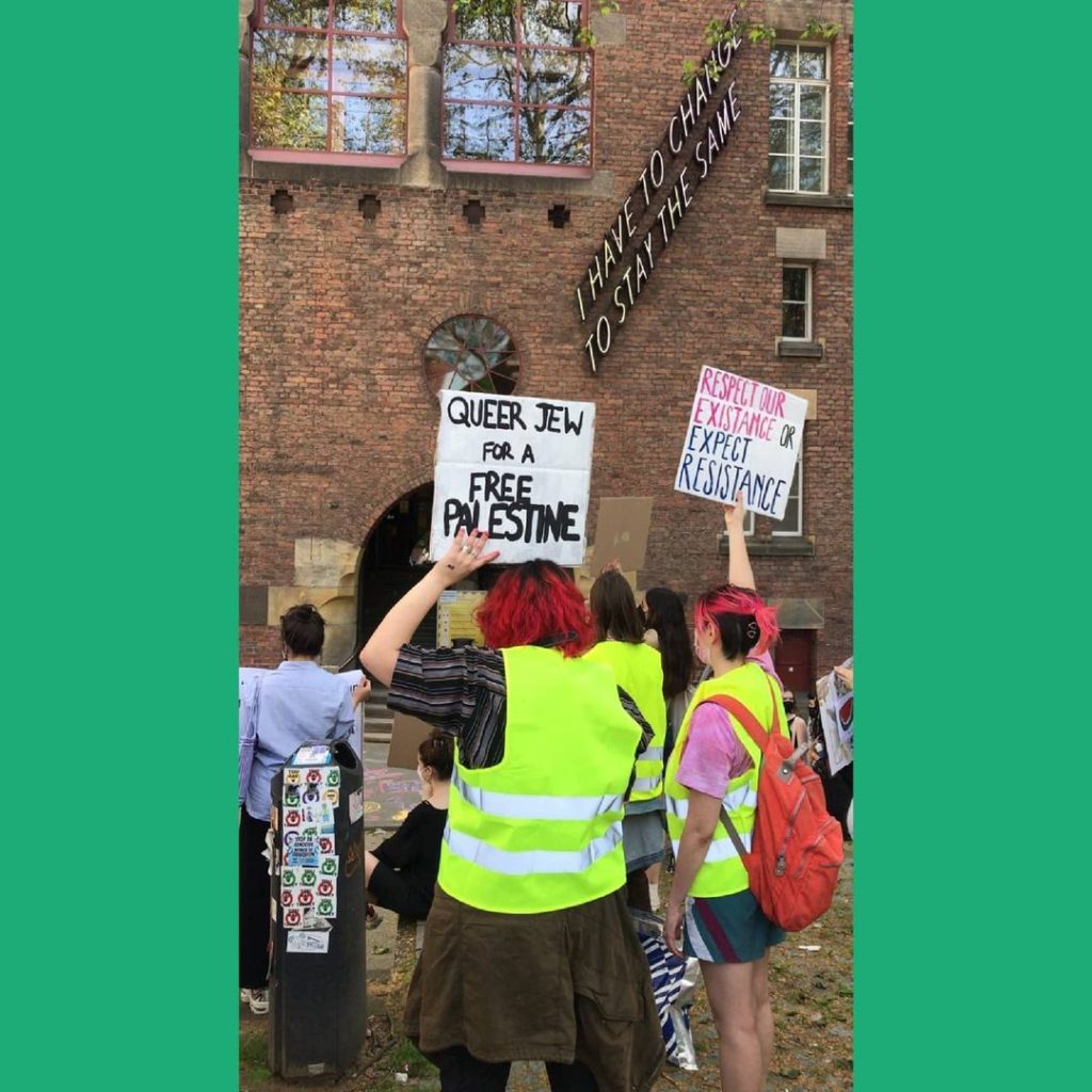 Demonstranten bij het schoolgebouw. Twee borden zijn zichtbaar, eentje met de tekst "Queer Jew for a free Palestine" en eentje met de tekst "Respect our existence or expect resistance".