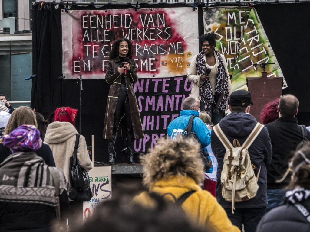 Het podium bij de demonstratie in Rotterdam. Kleurige spandoeken achter de sprekers. Er staan twee sprekers op het podium: Imani Gonsalves en Ramona Cairo, de moderators. Eén van hen lacht breeduit.