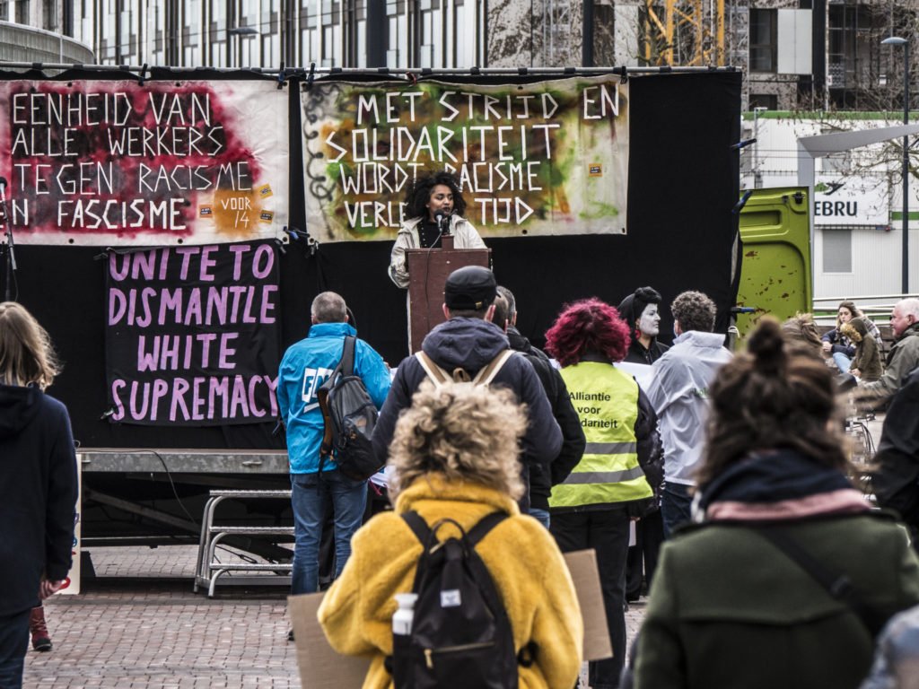 Een zwart persoon spreekt vanaf de lessenaar op het podium.