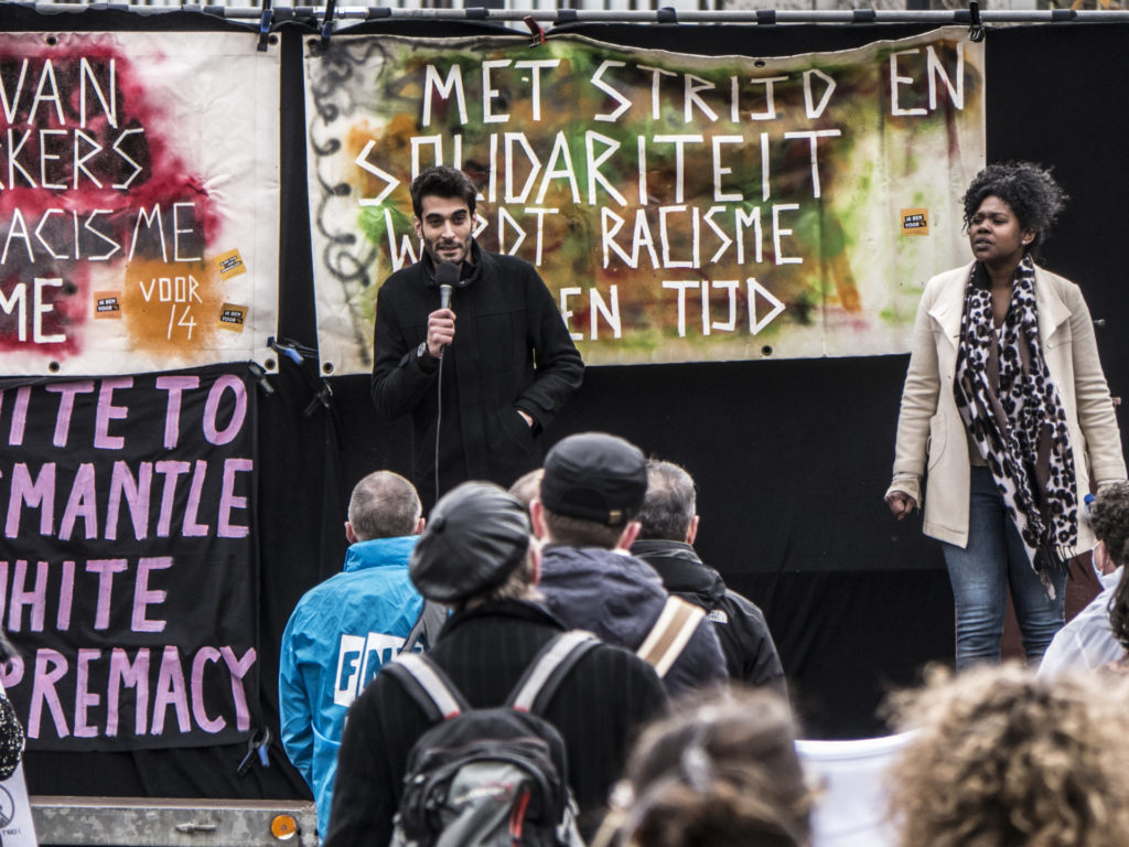 Eén van de sprekers (Ahmad al Sadeq) is aan het woord. Hij spreekt via een microfoon. Hij draagt een zwarte jas en heeft zwart, kort haar.