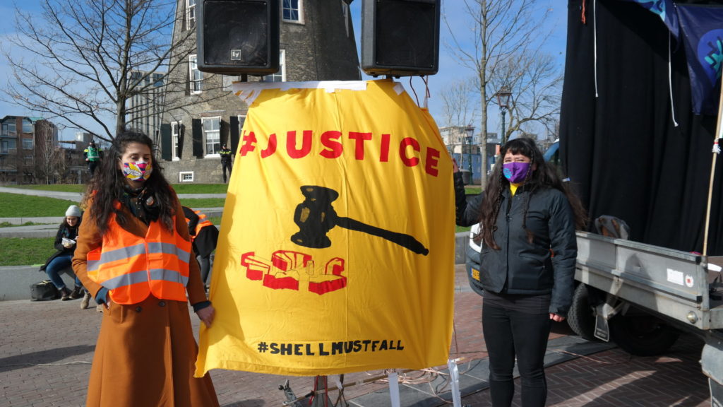 Een persoon met lang donker haar en een lange, wollen jas staat naast een groot geel spandoek met daarop de tekst "Justice - Shell must fall" en een afbeelding van een rechtershamer die het logo van Shell kapot slaat.